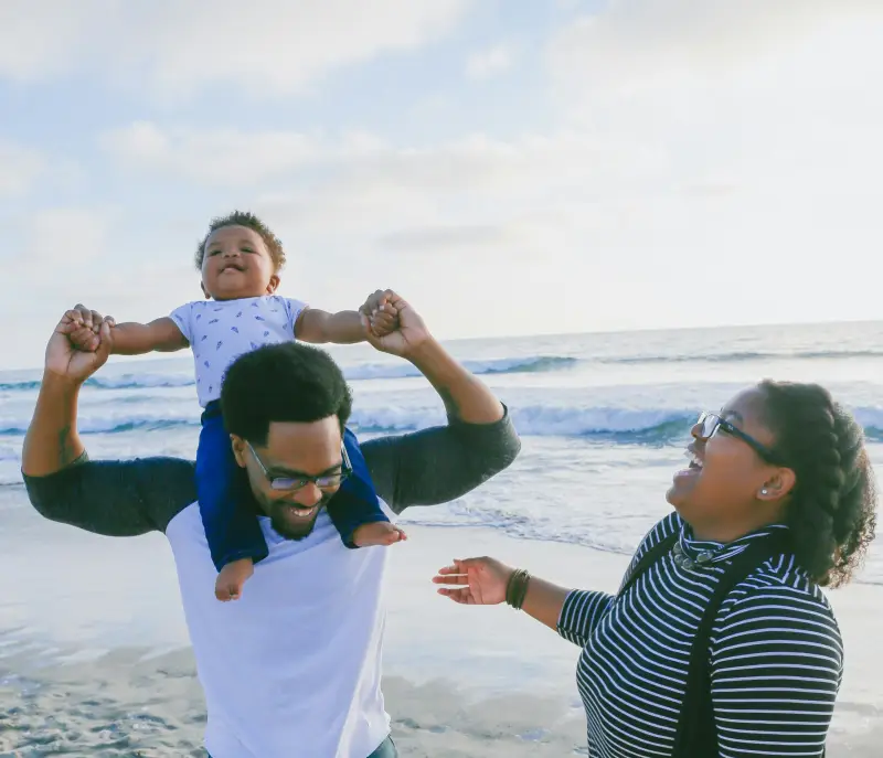 A father carries his baby on his shoulders at the beach, walking with his family.