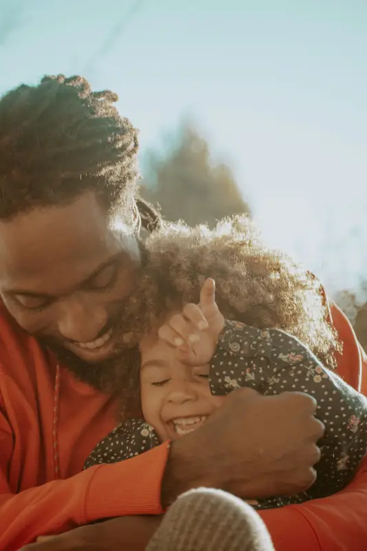 A father hugs his laughing young daughter in warm light.