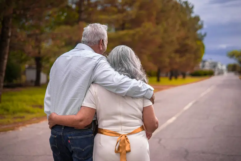 An older couple with grey hair walks arm in arm down a quiet road.