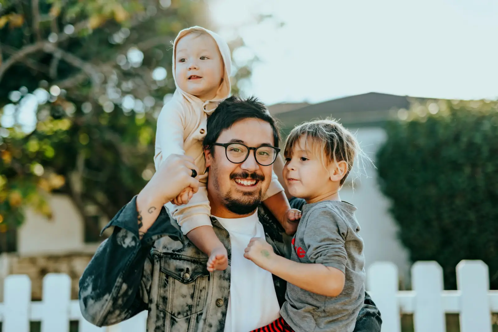 Smiling father carrying two young children in a backyard.