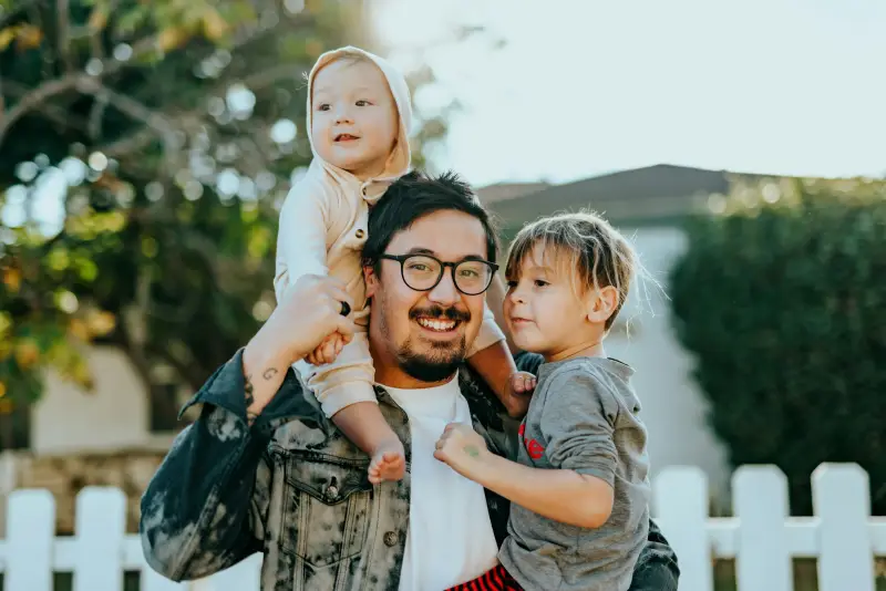 Smiling father carrying two young children in a backyard.